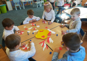 Grupa dzieci przy stoliku wykonuje kokardę narodową z biało czerwonych serc.